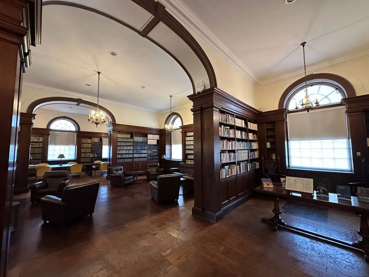 Interior of a traditional library with dark wooden bookshelves lining the walls and archways dividing the space. The room features large arched windows with white shades, chandeliers hanging from the ceiling, and several leather armchairs arranged for reading. A long wooden table with documents and display items sits near the front, and the shelves are filled with books.