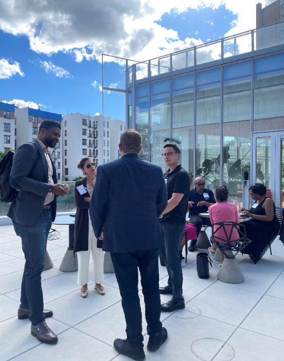 Four people standing and speaking, and three people sitting at a table eating and speaking on a rooftop patio.